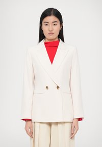 Dubbeltjesblazer in lichtcrème met gouden knopen, gestructureerde schouders en rechte mouwen, gedragen over een rood topje.