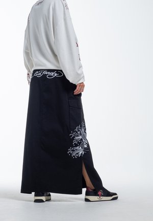 Long black skirt with side slit, featuring white fish and floral embroidery, paired with a white sweatshirt and black sneakers.