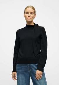 Czarny sweter z wysokim kołnierzem i detalem w postaci wiązania z boku, wykonany z miękkiego materiału. Posiada ribbrowane mankiety i dół. Połączony z niebieskimi dżinsami.