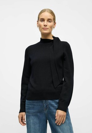 Czarny sweter z wysokim kołnierzem i detalem w postaci wiązania z boku, wykonany z miękkiego materiału. Posiada ribbrowane mankiety i dół. Połączony z niebieskimi dżinsami.