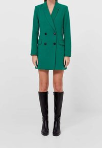 Vrouw draagt een groene double-breasted blazerjurk en kniehoge zwarte leren laarzen tegen een effen lichte achtergrond.