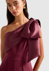Donna che indossa un abito senza maniche in satin bordeaux con un grande fiocco su una spalla e orecchini a cerchio dorati intrecciati.