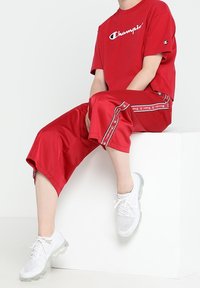 Czerona koszulka Champion z białym logo, dopasowane czerwone szorty z bocznym brandingiem oraz białe sneakersy sportowe o siatkowej fakturze.
