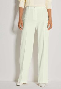 Vrouw in hoge taille, brede pijpen, lichtgekleurde broek met een crèmekleurige trui en witte puntige hakken, staande tegen een lichte muur.