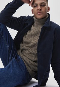 Jachetă din velur bleumarin peste un pulover cu guler înalt din tricot bej, asortată cu blugi de un albastru deschis. Modelul stă relaxat pe un fundal neutru.
