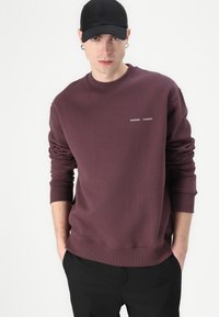 Sweatshirt bordeaux con polsini e scollatura a costine, con piccolo testo ricamato in bianco. Abbinato a pantaloni neri e un cappellino nero.