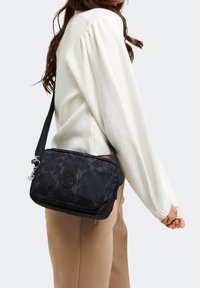 Sortmønstret crossbody taske lavet af tekstureret stof, med et logo badge og sølvfarvet hardware. Remmen kan justeres for komfort.