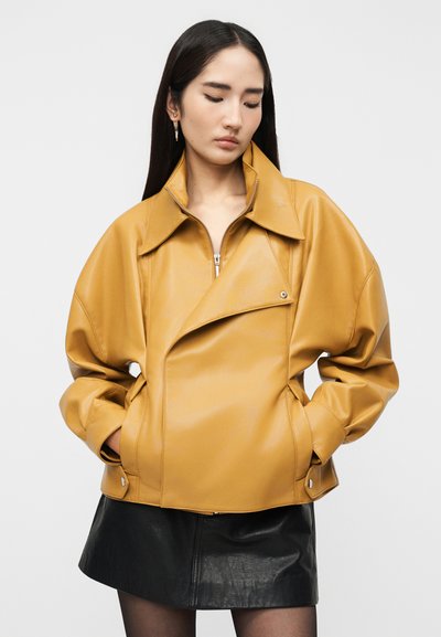 Γυναίκα με μακριά σκούρα μαλλιά, φορώντας ένα φαρδύ δερμάτινο μπουφάν μουσταρδί και μια μαύρη δερμάτινη φούστα, με τα χέρια στις τσέπες, κοιτάζοντας προς τα κάτω.