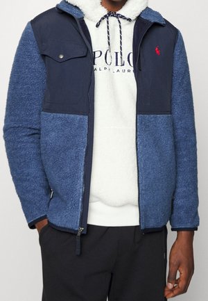 Hombre vistiendo chaqueta de forro polar azul sobre una sudadera con capucha blanca Polo Ralph Lauren, con las manos parcialmente en los bolsillos, frente a un fondo liso.