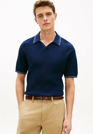 Junger Mann in navyblauem, strukturiertem Kurzarm-Poloshirt und beiger Hose mit braunem Gürtel steht mit den Händen in den Taschen vor weißem Hintergrund.