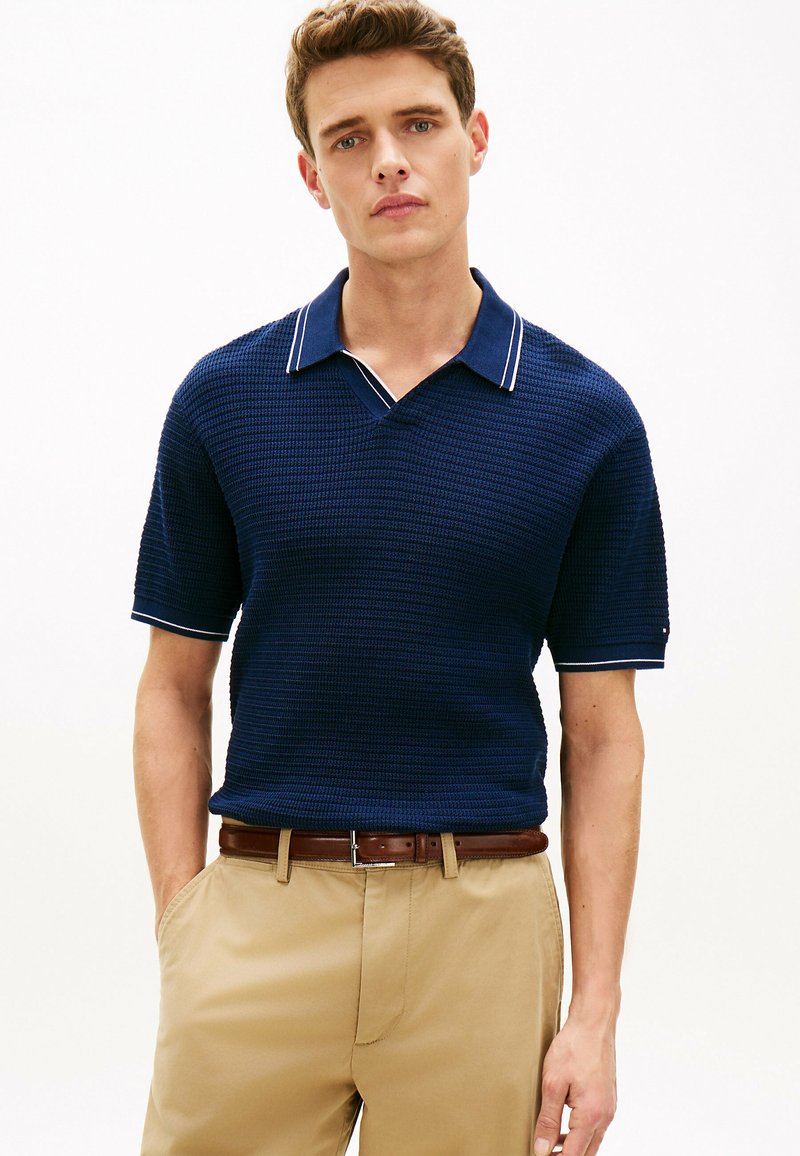 Jeune homme portant un polo bleu marine texturé à manches courtes et un pantalon beige avec une ceinture marron, debout les mains dans les poches devant un fond blanc.
