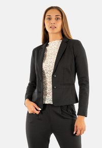 Blazer noir ajusté en tissu texturé, à boutonnage simple, avec des revers à cran. Porté sur un haut à motif léger, associé à un pantalon assorti.