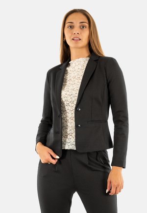 Blazer noir ajusté en tissu texturé, à boutonnage simple, avec des revers à cran. Porté sur un haut à motif léger, associé à un pantalon assorti.