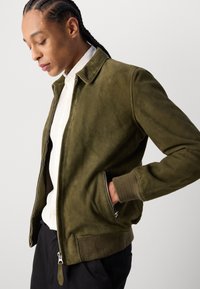 Giacca in suede verde oliva con zip frontale, polsini a costine e colletto; presenta tasche laterali e una texture morbida. Indossata sopra una camicia bianca.