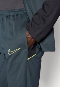 Športové nohavice Nike v tmavom tealovom odtieni s čiernym pásom, bočnými vreckami s žltým lemovaním a malým bielym logom.