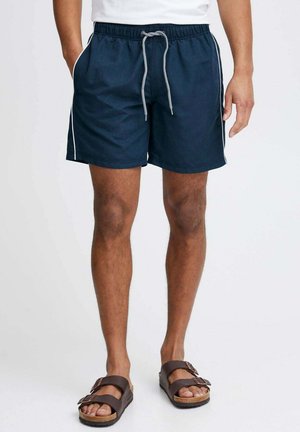 Pantaloni da nuoto blu navy con vita regolabile con cordino, strisce laterali bianche e tasche. Abbinati a sandali marroni con doppie cinghie.