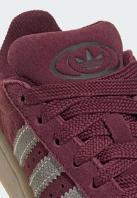 Gros plan d'une basket en suède bordeaux avec des lacets texturés, des bandes grises et un logo Adidas noir sur la languette rembourrée.