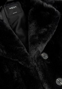 Manteau en fausse fourrure noire avec une texture douce, un large col et deux fermetures à bouton. L'étiquette intérieure affiche la marque "NEXT."