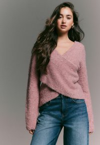 Giovane donna con lunghi capelli scuri che indossa un maglione avvolgente rosa e jeans blu, in piedi contro uno sfondo grigio semplice.