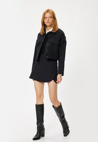 Veste noire courte avec deux poches avant et des boutons-pression, portée sur une robe noire ajustée. Le mannequin porte des bottes noires jusqu'aux genoux.