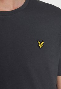 Tmavě šedé tričko s krátkým rukávem vyrobené z měkké bavlny. Má vyšitý žlutý logotyp orla na hrudi. Kulatý výstřih.