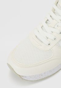 Zapatilla blanca con parte superior de malla, acentos de cuero liso, punta redonda, cordones tonales y una suela texturada con manchas.