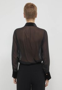 Schwarzes, transparentes Langarmhemd mit Kragen und Samtabnähern, das aus einem strukturierten Stoff gefertigt ist. Figurbetonter Schnitt mit abgerundetem Saum und Knopfakzenten.