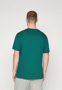 Camiseta de algodón verde esmeralda con mangas cortas y cuello redondo. La parte de atrás es lisa y tiene un corte relajado, combinada con pantalones gris claro.
