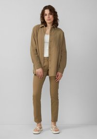 Jasnobrązowa koszula na guziki z teksturowanej tkaniny, noszona na białym topie na ramiączkach, w zestawie z beżowymi spodniami i białymi sandałami na paskach.