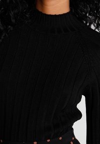 Schwarzer gerippter Strickpullover mit hohem Kragen und Raglanärmeln, der ein strukturiertes Muster aufweist. Der Stoff wirkt weich und flexibel.
