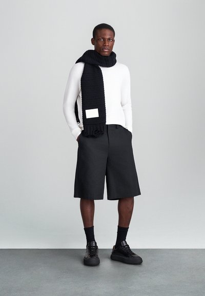 Mustro mustriga sall, valge pika varrukaga särk, mustad lühikesed püksid ja mustad jooksukingad. Erakordne ruudukujuline silt sallil.