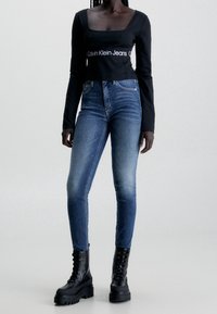 Svart långärmad topp med fyrkantig halsringning och vit logodetalj, ihop med smala blå jeans och svarta klumpiga snörstövlar.