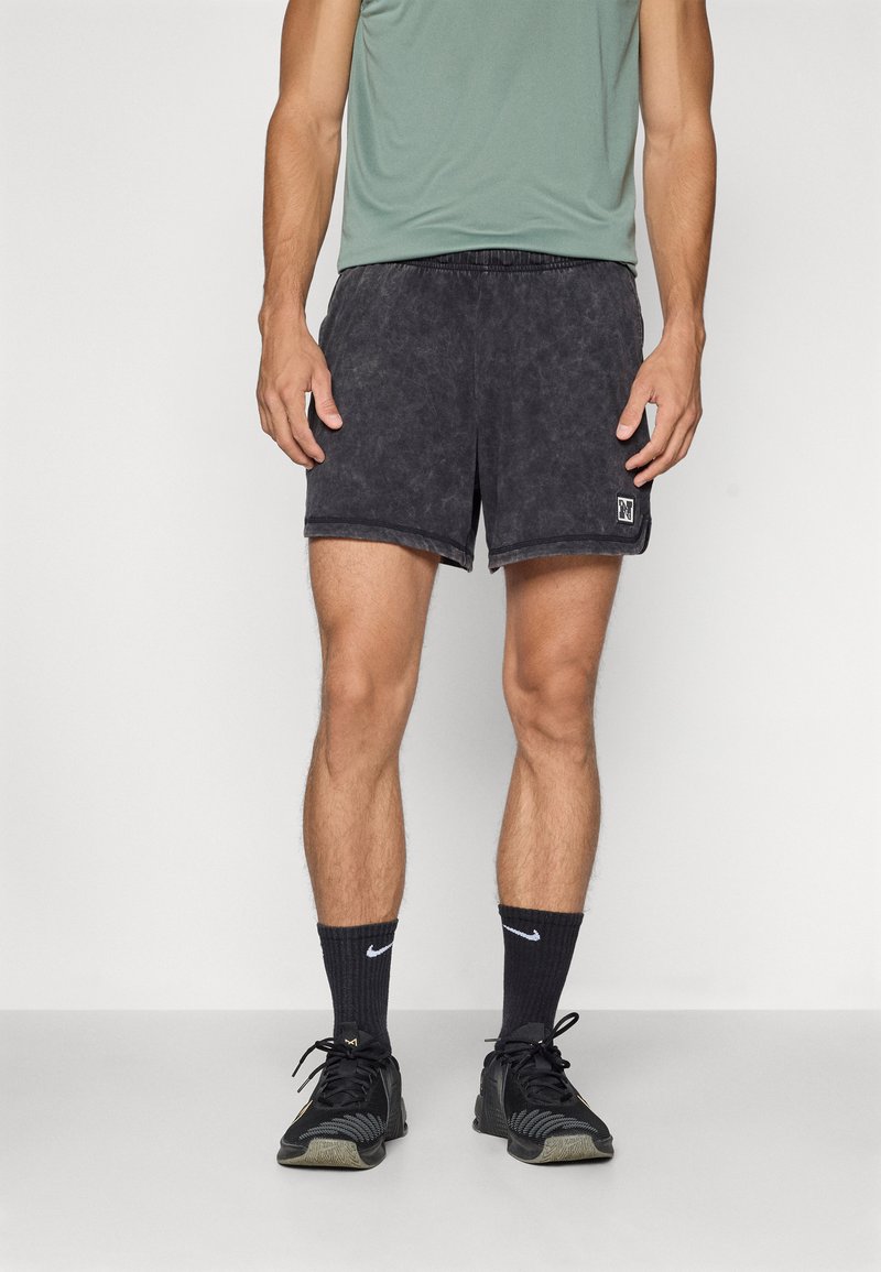 Sorte, teksturerede shorts med elastisk talje, parret med en grøn skjorte og sorte træningssko, stående foran en hvid baggrund.