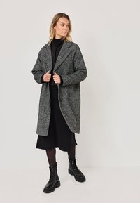 Pelēks teksturēts mētelis ar brīvu piegriezumu un lieliem atlokiem, uzlikts virs melna apģērba, kopā ar melniem kurpēm ar šņorēm.