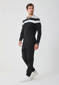 Schwarzes langärmeliges Polohemd mit grauem Kragen und weißen horizontalen Streifen. Kombiniert mit schwarzen Cargo-Hosen und weißen Turnschuhen.