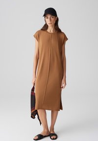 Braunes Midikleid mit kurzen Ärmeln, Mittelnaht, seitlicher Schlitze und glatter Textur. Kombiniert mit einer schwarzen Kappe und Rutschsandalen, während ein gemusterter Schal gehalten wird.