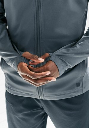 Gray zip-up jacket with fitted cuffs, smooth texture, and a structured design. Hands are positioned in front, partially obscured by sleeves.