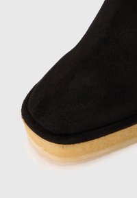 Parte superior de zapato de ante negro con puntera redondeada y suela gruesa de goma crepe beige sobre fondo blanco.