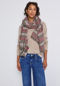 Vævet tørklæde i beige, brun og rød hundetand mønster, med frynser i kanten, båret over en beige sweater og blå denim jeans.
