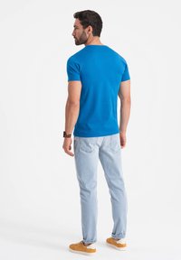 T-shirt di cotone blu con maniche corte, indossata con jeans chiari e sneakers beige, vista da dietro, che mette in evidenza la vestibilità e il colore.