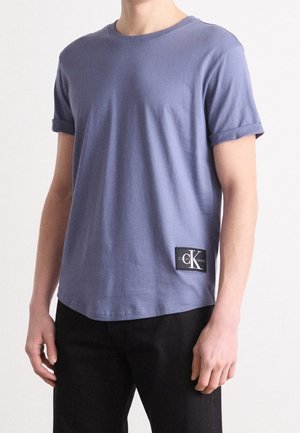T-shirt à manches courtes et col rond en coton lavande clair avec manches retroussées, portant un patch logo Calvin Klein noir en bas à gauche.
