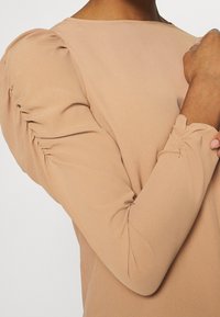 Maglia a maniche lunghe in morbido tessuto beige con spalle a sbuffo, presenta dettagli arricciati ai polsini delle maniche e una texture liscia.