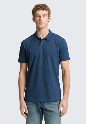 Młody mężczyzna o kręconych włosach, ubrany w granatową koszulkę polo z krótkim rękawem i jasnoniebieskie dżinsy, stoi na jednolitym jasnym tle.