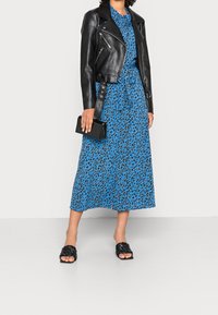 Czarna skórzana kurtka motocyklowa nałożona na midi sukienkę w niebieski wzór panterki. Czarna torebka kopertowa i czarne plecione sandały dopełniają stylizację.