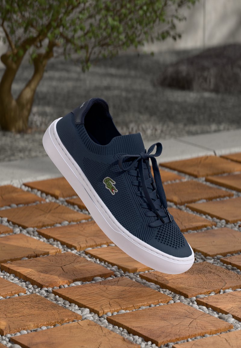 Sötétkék kötött sneaker fehér talppal, texturált felsőrész és fűzős zárás. Oldalán zöld logó található, amely fából és kavicsokból álló háttér előtt látható.