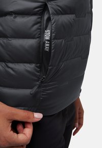 Schwarze Puffer-Weste mit gestepptem Design, ausgestattet mit einer Reißverschlusstasche, die mit "STOW AWAY" beschriftet ist. Glatte Textur mit leichtem Erscheinungsbild.