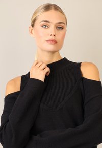 Czarny dziany sweter z wysokim kołnierzem i otwartymi ramionami. Tekstura jest miękka, z prążkowanymi detalami przy kołnierzu i ramionach.