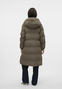 Manteau long rembourré en vert olive avec une capuche, présentant une texture matelassée et des poignets élastiques. Porté sur un jean foncé et des chaussures beiges.