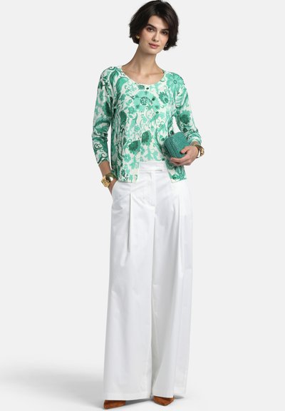 Sieviete valkā zaļu ziedu raksta garroku topu, baltas platajam kāju bikses, brūnus apavus, zelta aproces, tur tirkīza krāsas rokassomiņu, stāvot pret baltu fonu.