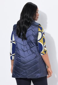 Marineblau gesteppte Weste mit Zickzackmuster, ausgestattet mit einem hohen Kragen und leichtem Material. Die Rückansicht zeigt eine glatte Textur und ein ärmelloses Design.
